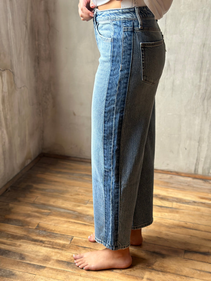 A person wearing wide-leg cropped denim jeans with a unique dual-panel design, standing barefoot on a wooden floor against a textured concrete wall.