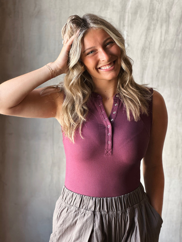 Woman wearing a purple sleeveless top and gray pants against a neutral background