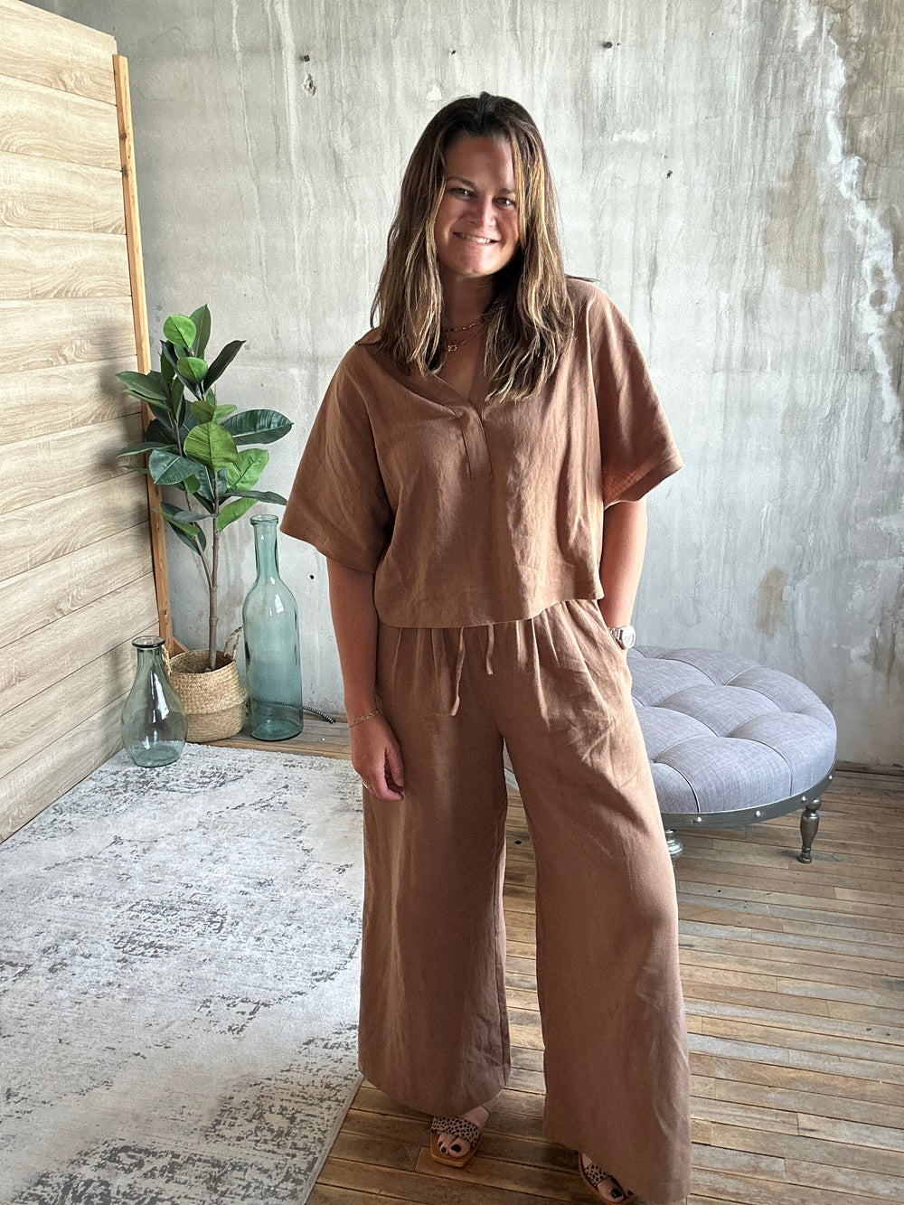 Person wearing a brown outfit standing in a room with a plant and a chair.