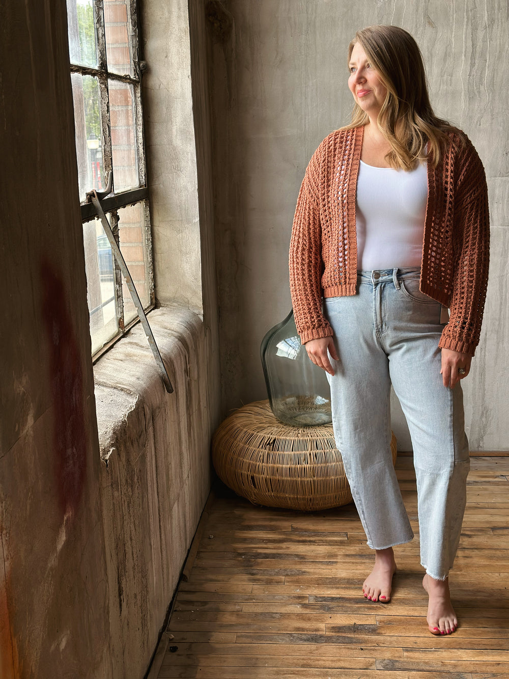 A woman standing in a room with a wooden floor and window, wearing a light brown knitted cardigan.