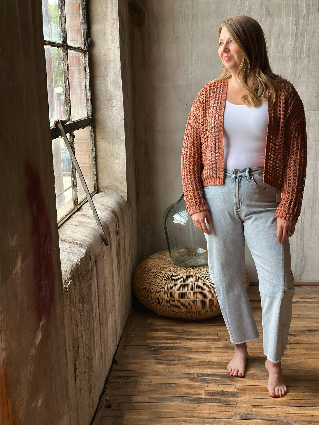 A woman standing in a room with a wooden floor and window, wearing a light brown knitted cardigan.