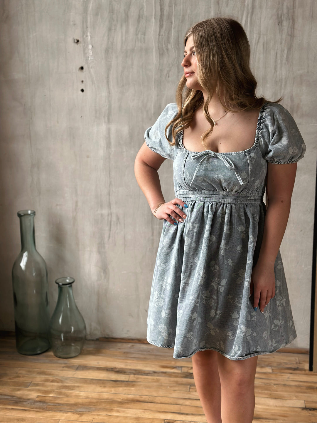 Woman wearing a light blue denim floral dress standing against a textured wall.