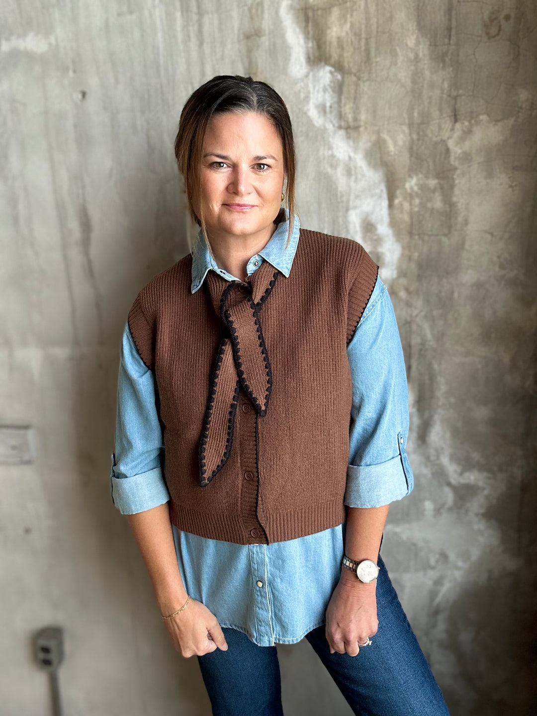 A woman wearing a brown ribbed knit vest with a black braided tie detail over a light blue button-down shirt and blue jeans, standing against a textured concrete wall.