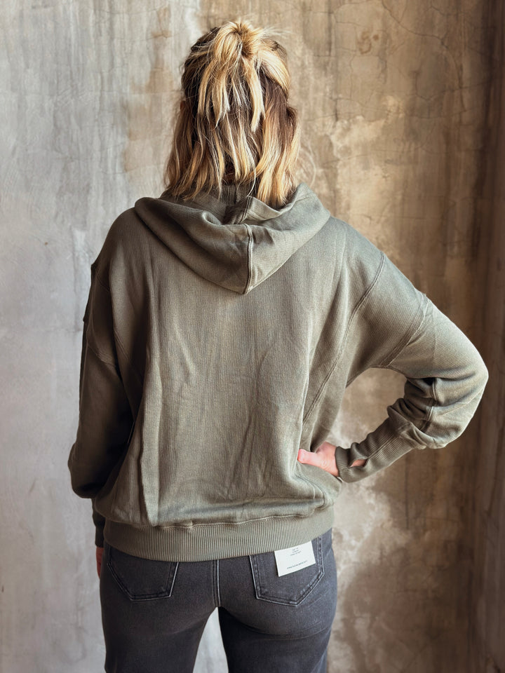 Person wearing a green hoodie and dark jeans against a textured wall.