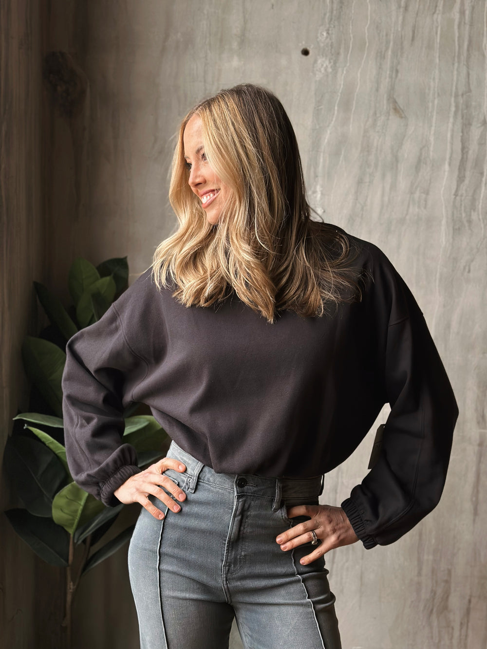 Woman wearing a dark gray sweater and light gray jeans against a concrete wall.