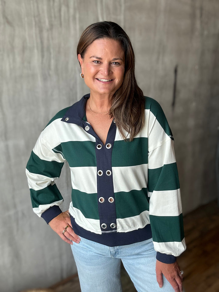 A woman wearing a green and white striped long sleeve with a navy blue collar, cuffs, and hem, paired with light blue jeans, standing against a textured gray wall.