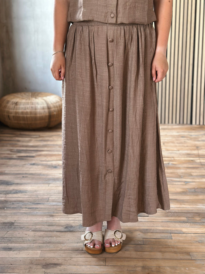A person wearing a brown skirt with buttons standing on a wooden floor.