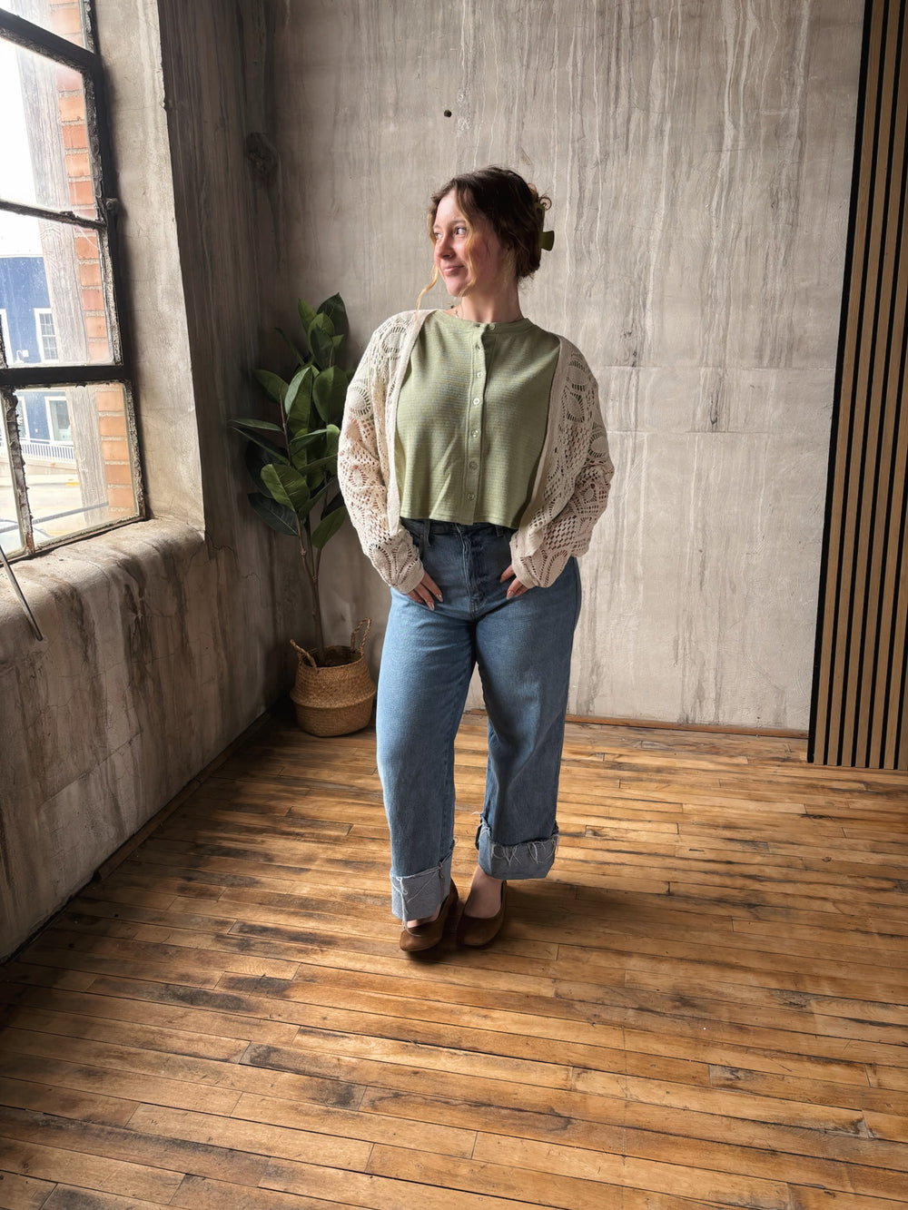 Woman standing in a room with wooden floor and concrete wall, wearing a light green blouse and blue jeans.