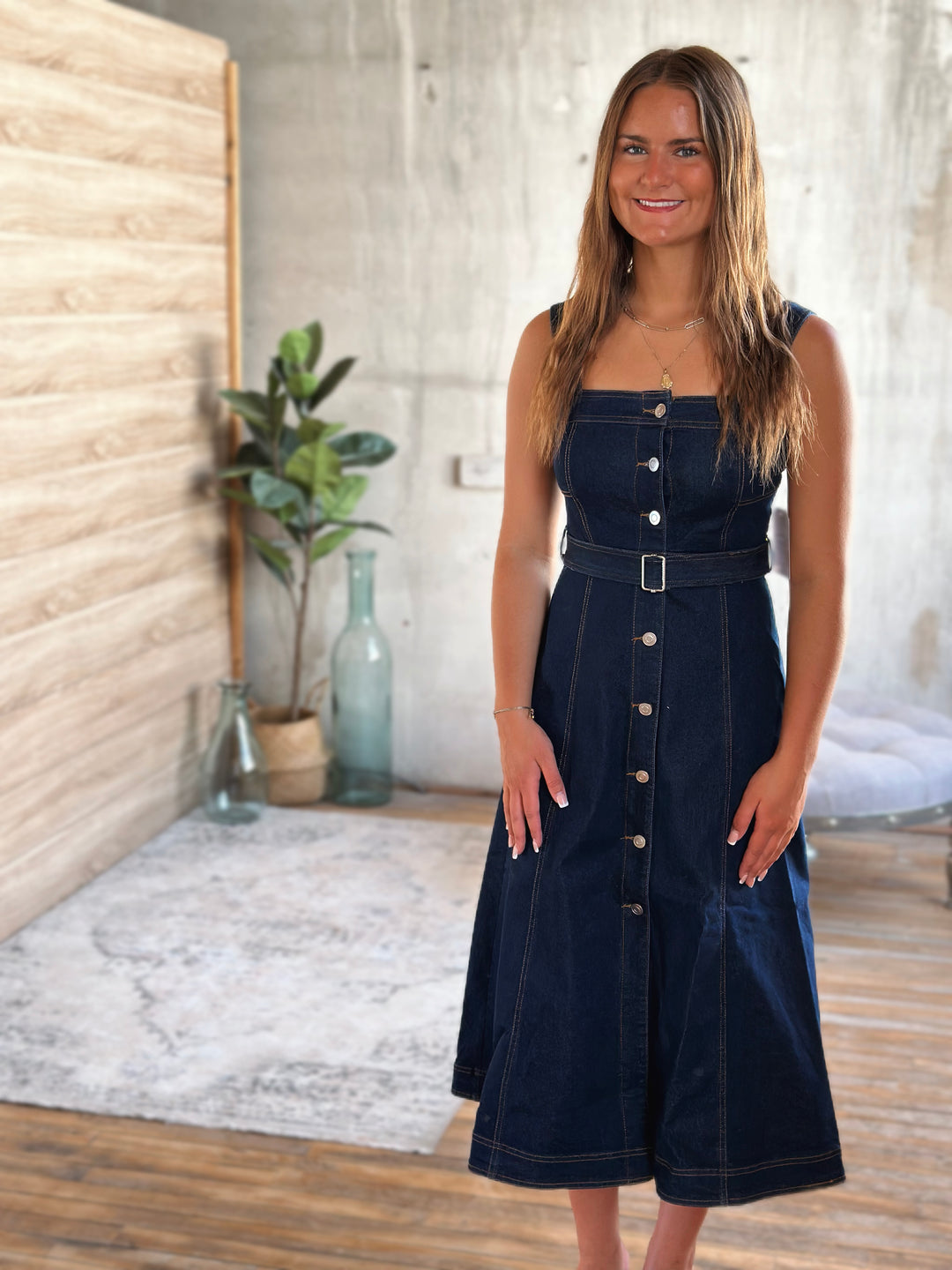 Woman wearing a dark blue denim dress with silver buttons in a room with wooden floor and plant.