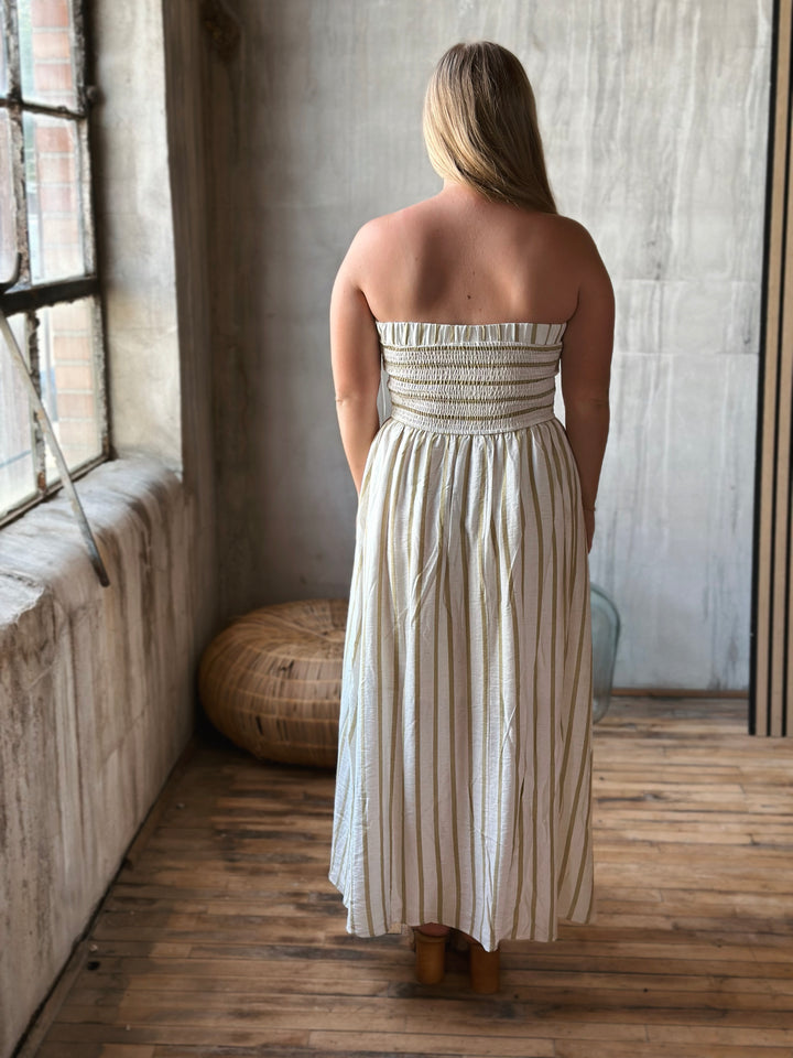 Woman wearing a long white strapless dress with beige stripes in a room with wooden floor and textured walls.