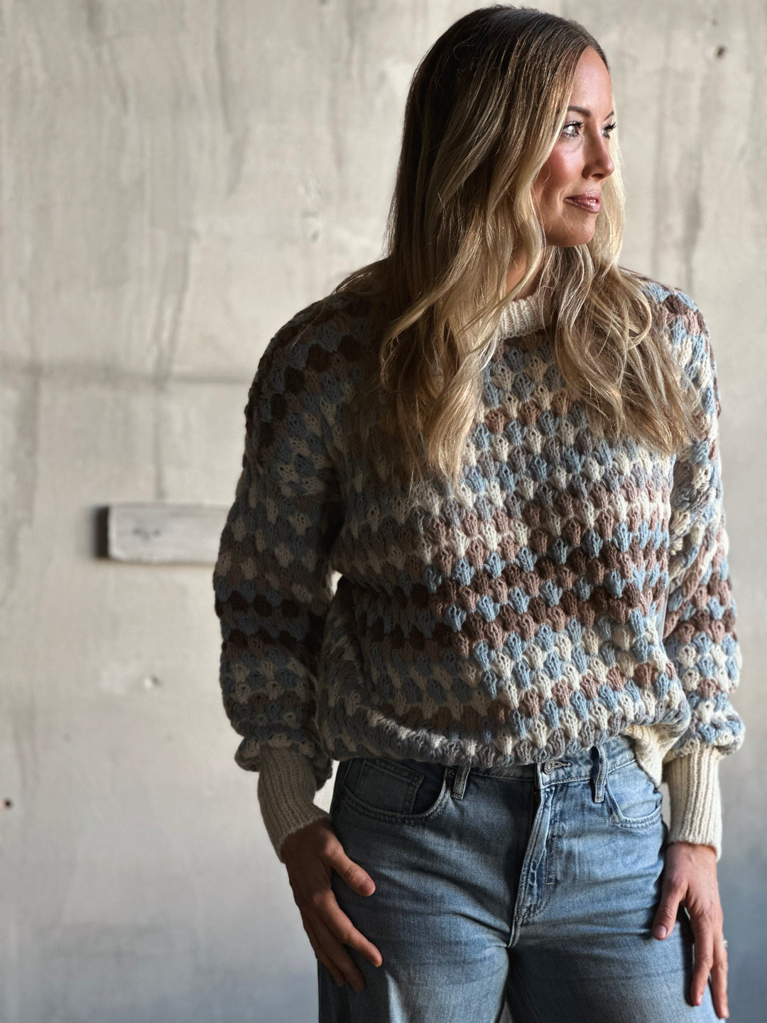 Woman wearing a patterned sweater and jeans against a concrete wall.