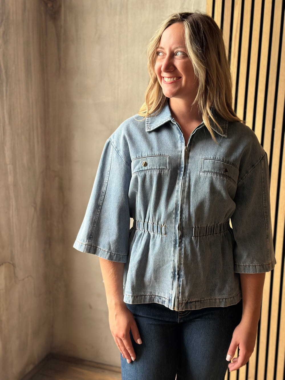 A woman wearing a light blue denim cropped button-up shirt with three-quarter sleeves and dark blue jeans, standing in front of a textured concrete wall and a slatted wood panel background.