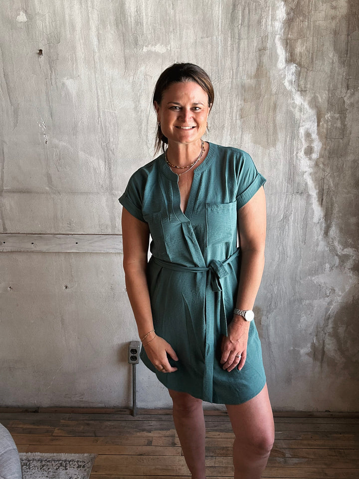 Woman wearing a dark teal dress with strings to tie in the front and pockets  standing against a textured wall.