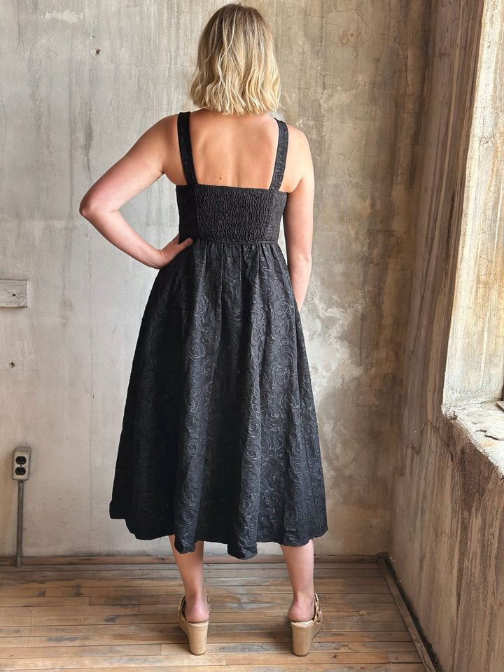 Woman wearing a black dress standing in a room with wooden floor and concrete wall.