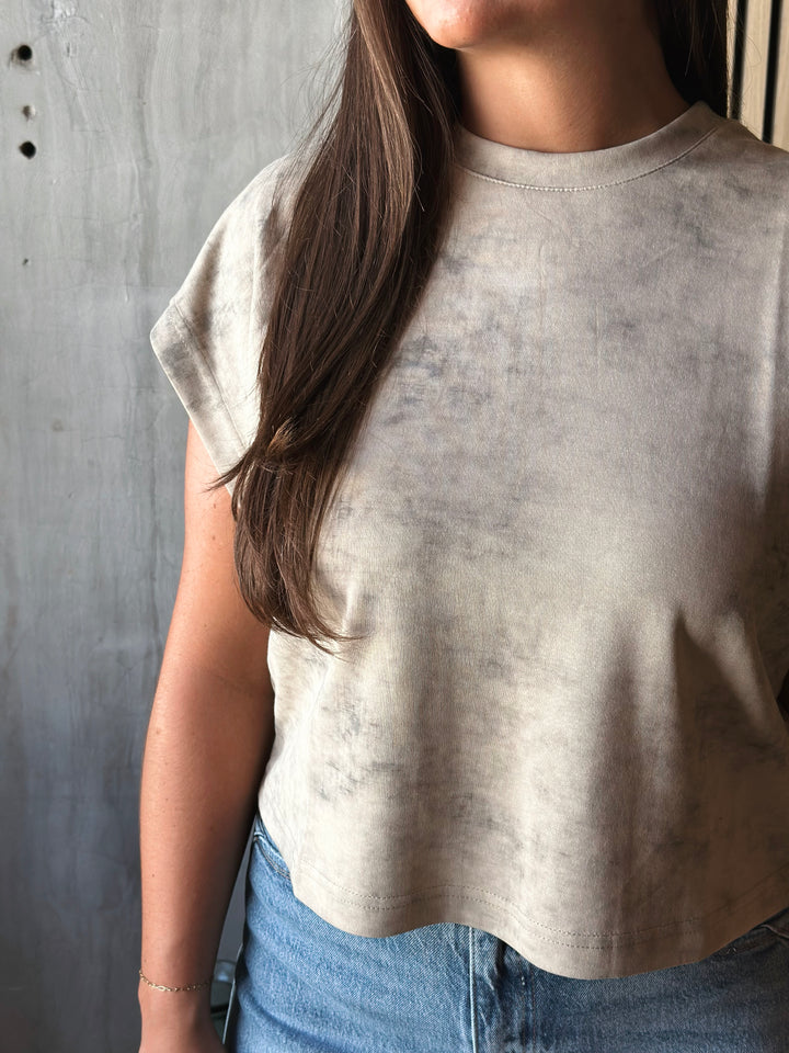 Person wearing a beige t-shirt and blue jeans against a gray wall