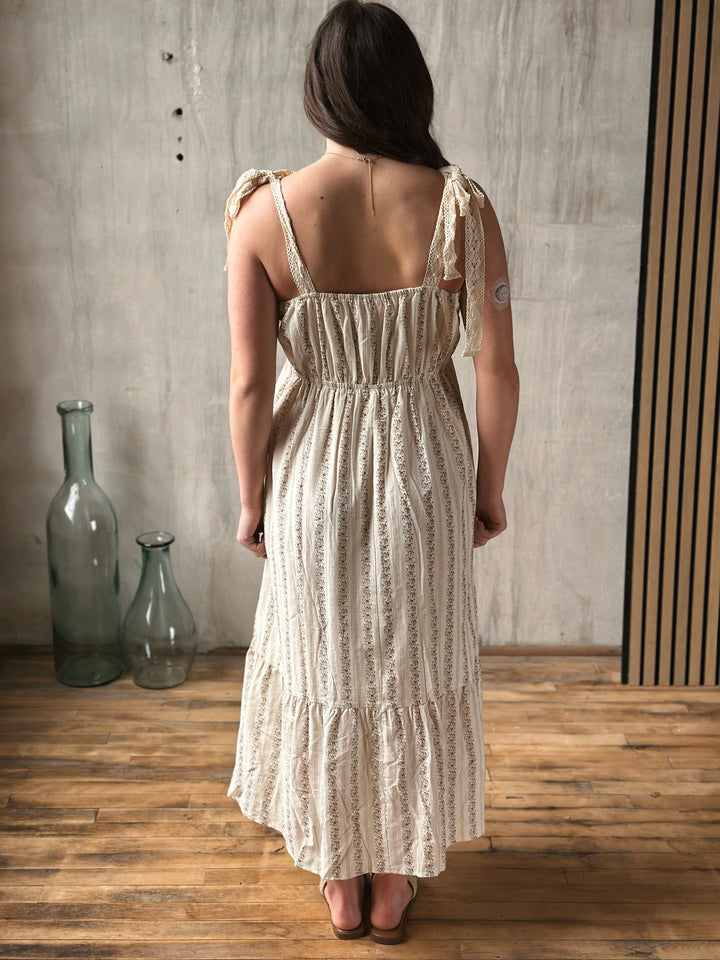 Woman wearing a long, light-colored dress with thin straps in a room with wooden floor and gray wall.
