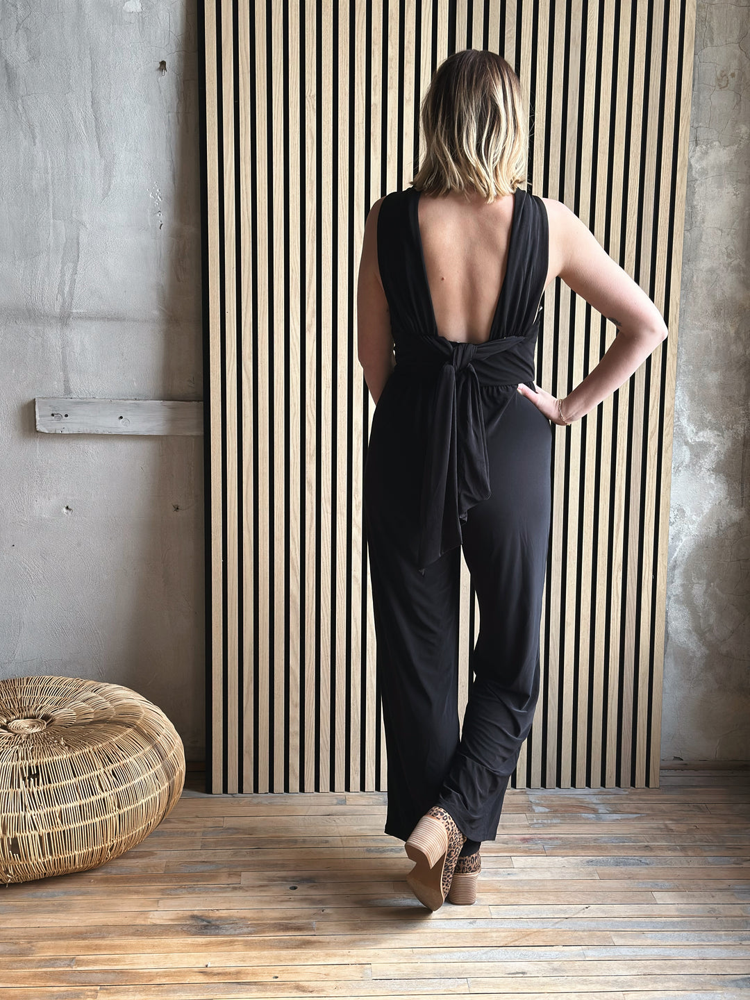 Woman wearing a black jumpsuit with an open back in a room with wooden flooring and a textured wall.