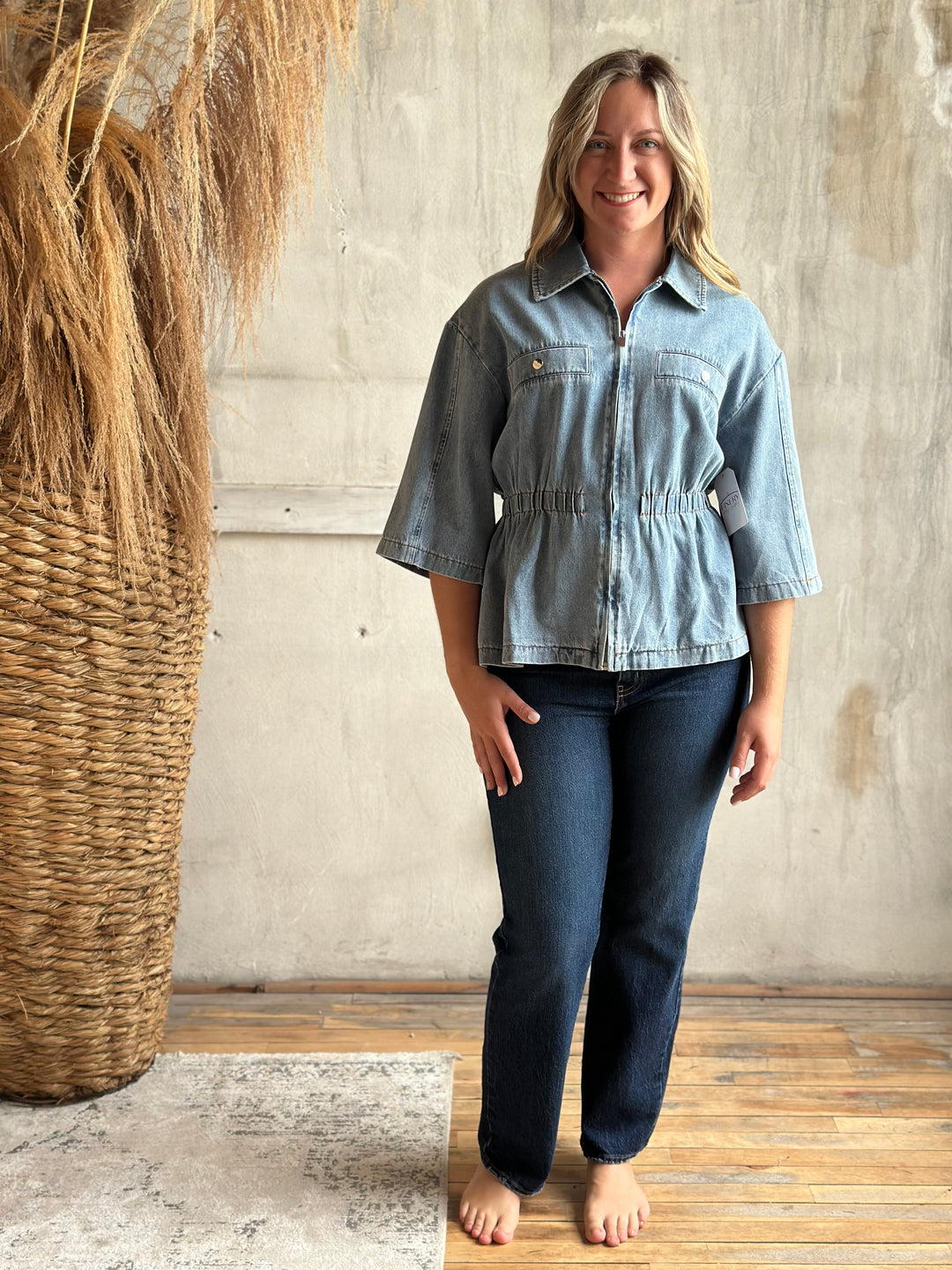 A woman stands barefoot in a rustic indoor setting, wearing a light blue cropped denim jacket with three-quarter sleeves and dark blue jeans.