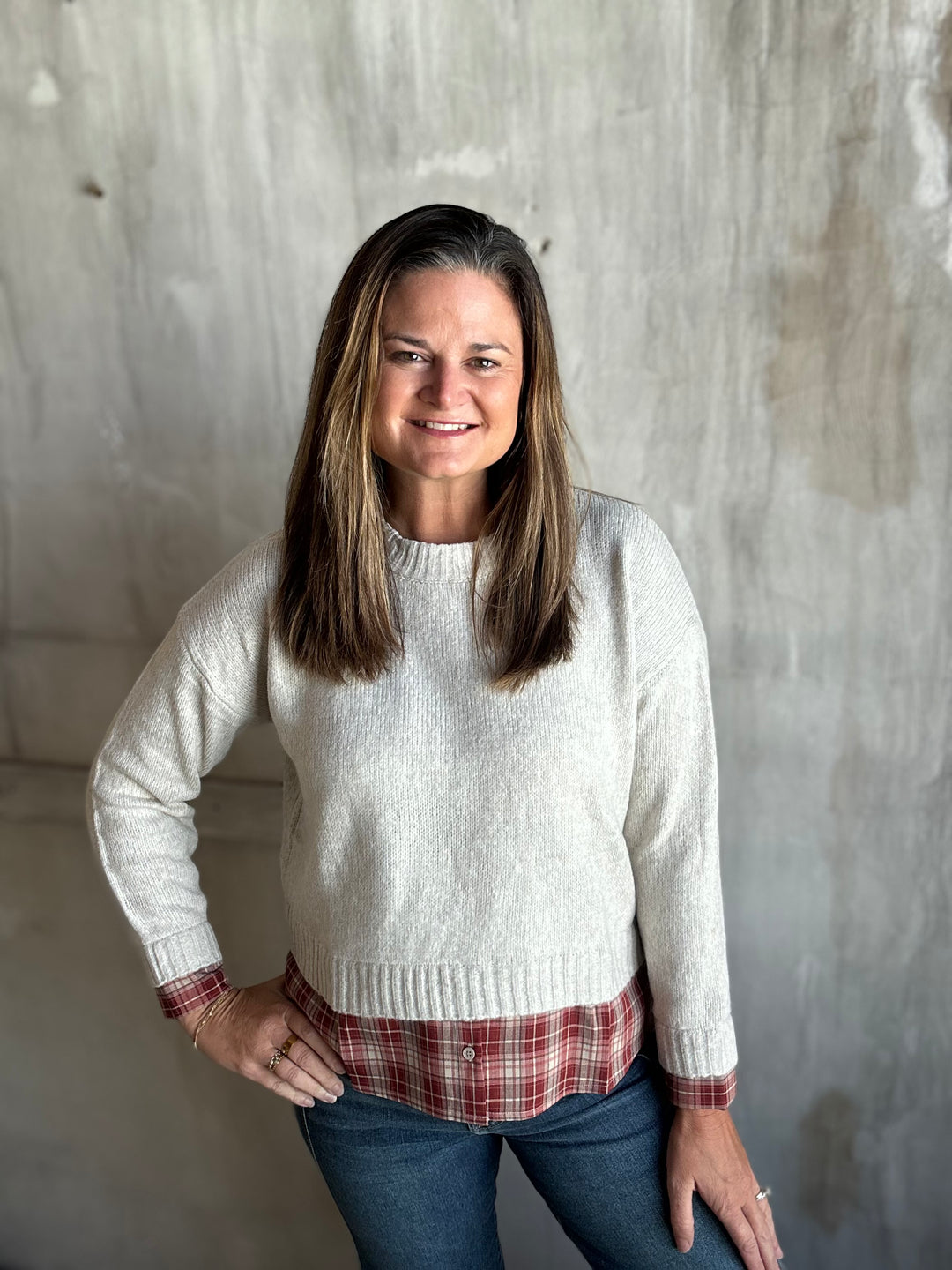 A woman with shoulder-length brown hair stands against a textured concrete wall, wearing a cream-colored knit crewneck sweater over a red and white plaid shirt, paired with blue jeans.