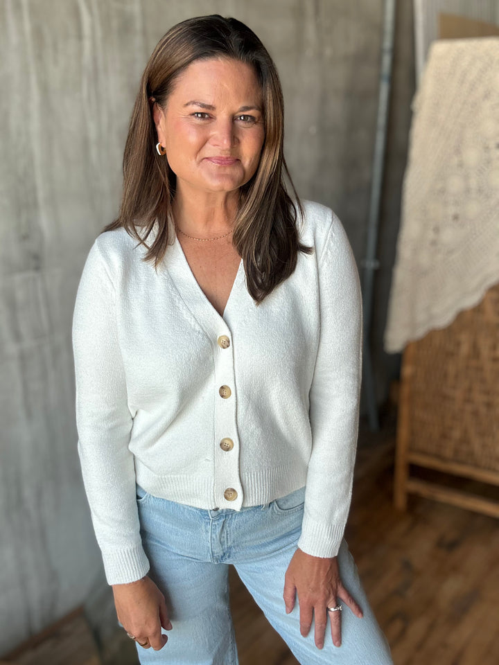 A woman wearing a white knit cardigan with brown buttons and light blue jeans.