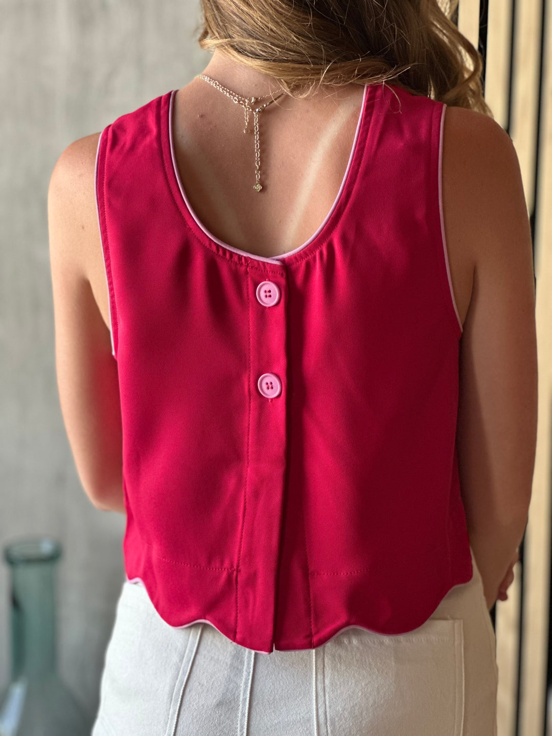 Back view of model wearing a scarlet colored top with a sleeveless design and scalloped hem and a button closure on the back of top