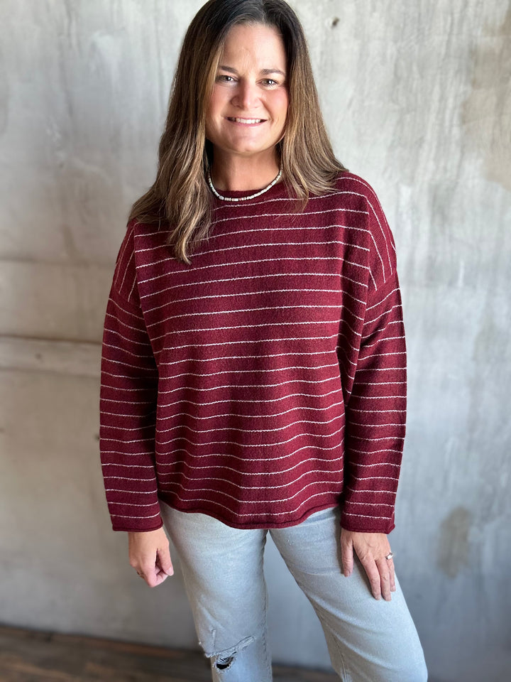 A woman wearing a long-sleeve sweater with thin horizontal burgundy and white stripes, paired with light gray jeans, standing against a textured concrete wall.