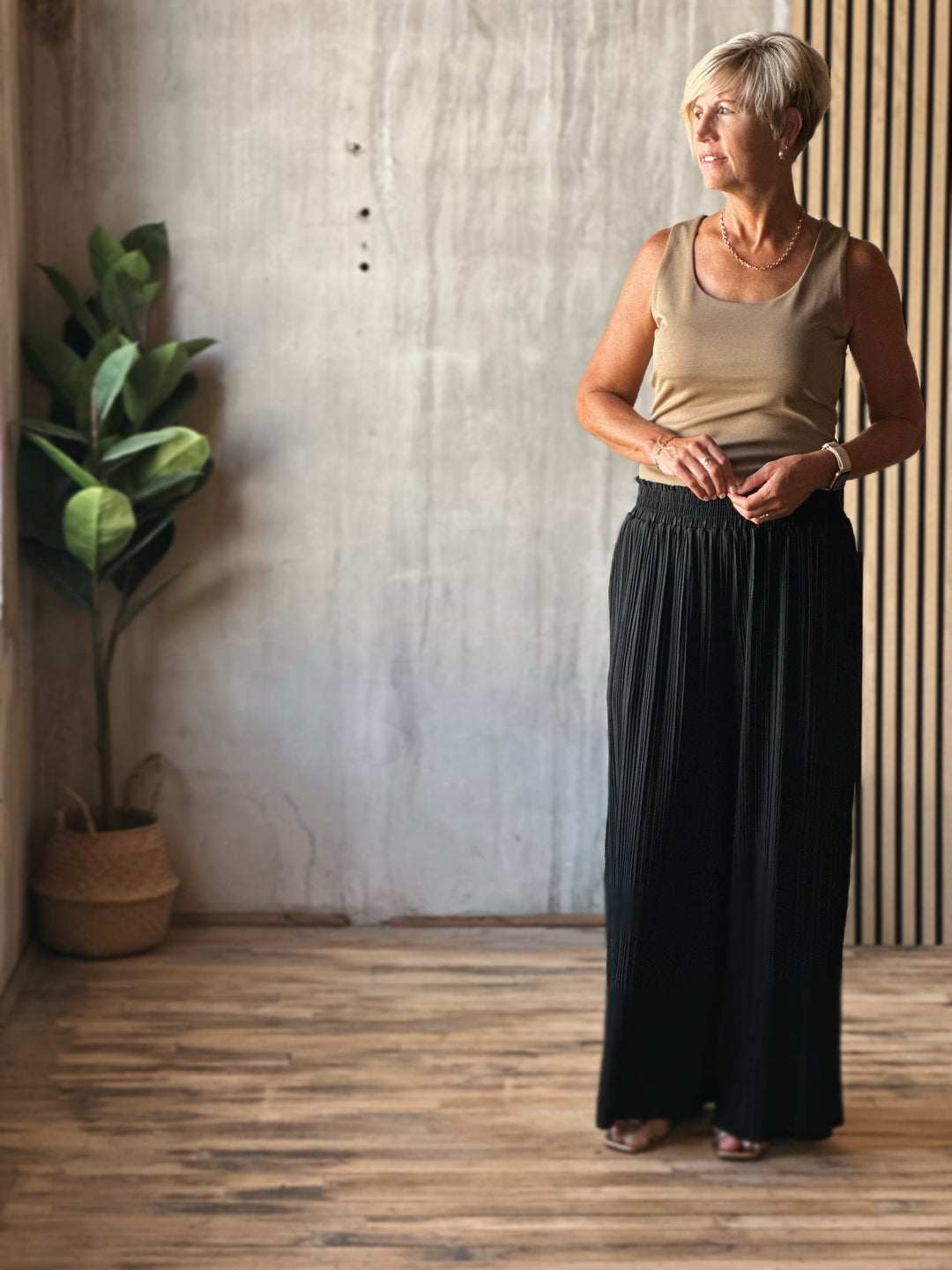 Woman standing in a room with wooden floor and plant, wearing a beige top and black skirt.