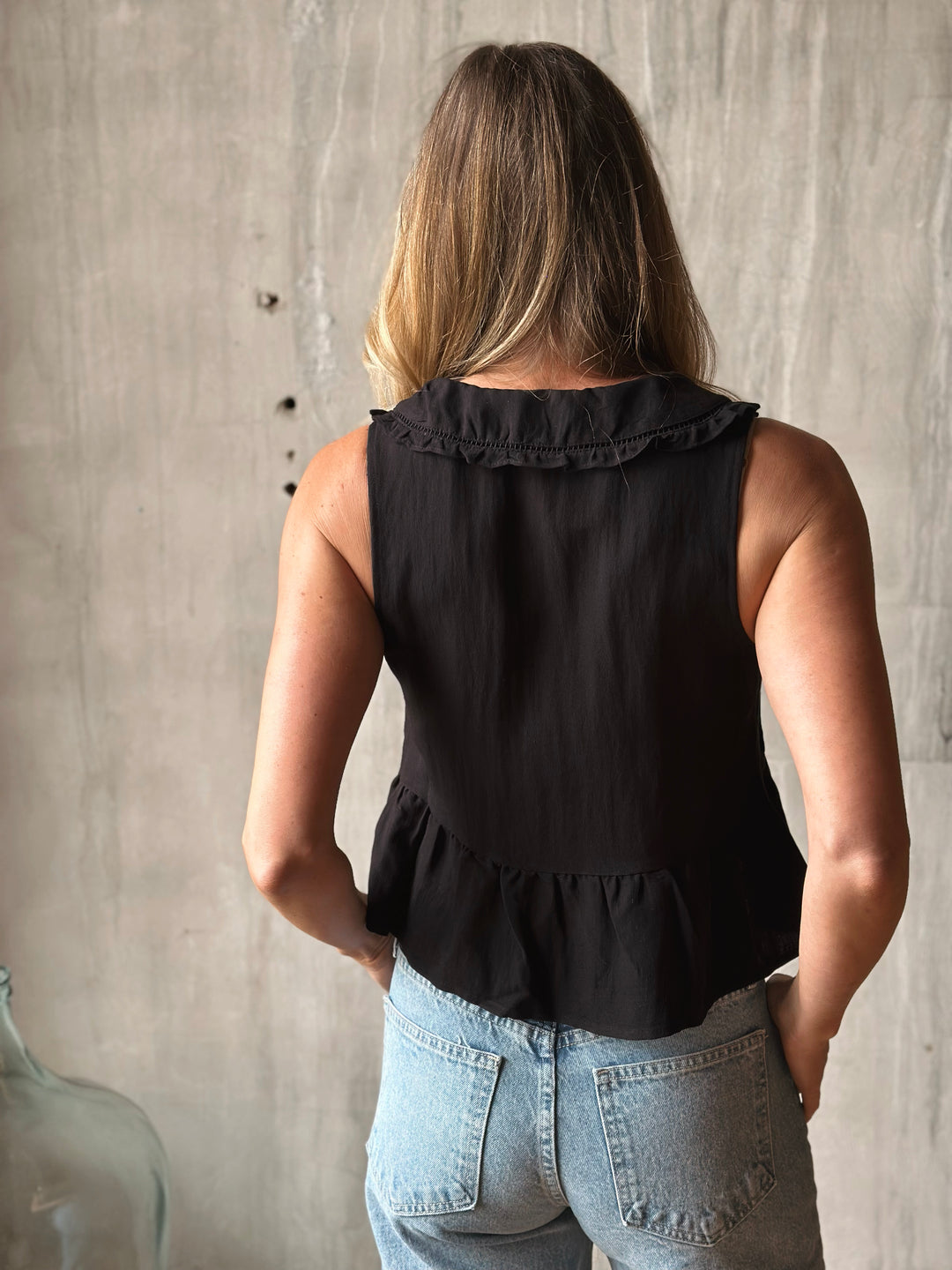 Person wearing a black sleeveless top with ruffled details against a neutral wall.