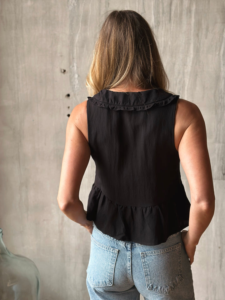 Person wearing a black sleeveless top with ruffled details against a neutral wall.