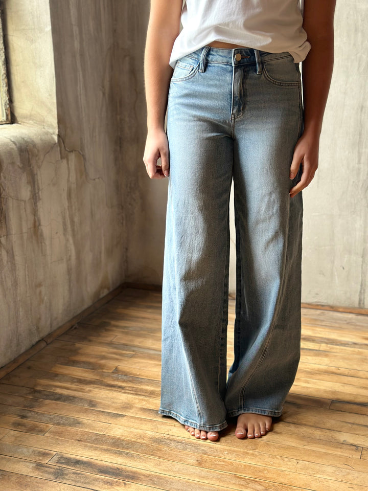 A person wearing light-wash wide-leg denim jeans and a white top, standing barefoot on a wooden floor against a textured concrete wall.