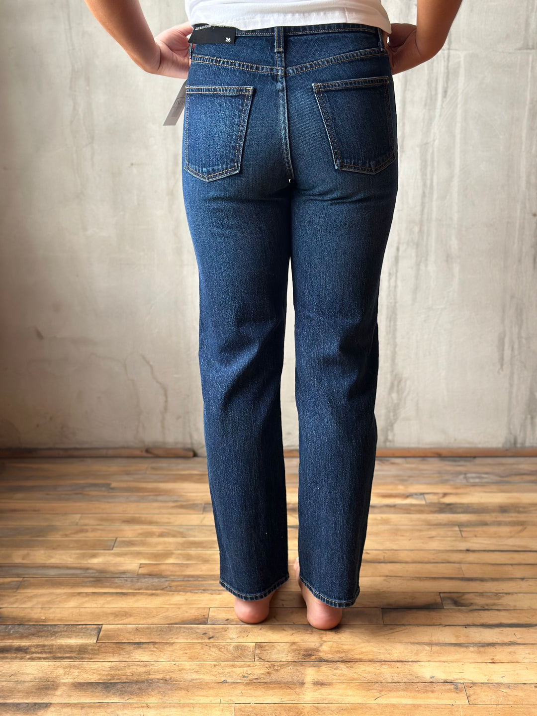 A person shown from the back wearing dark blue straight-leg jeans, standing on a wooden floor against a textured concrete wall.