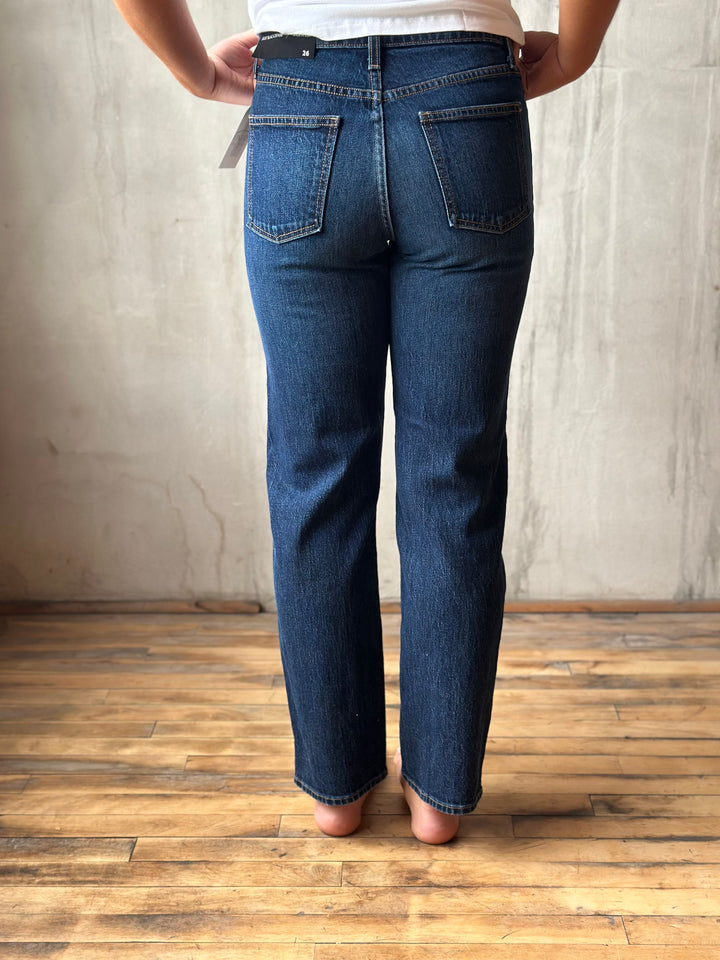 A person shown from the back wearing dark blue straight-leg jeans, standing on a wooden floor against a textured concrete wall.