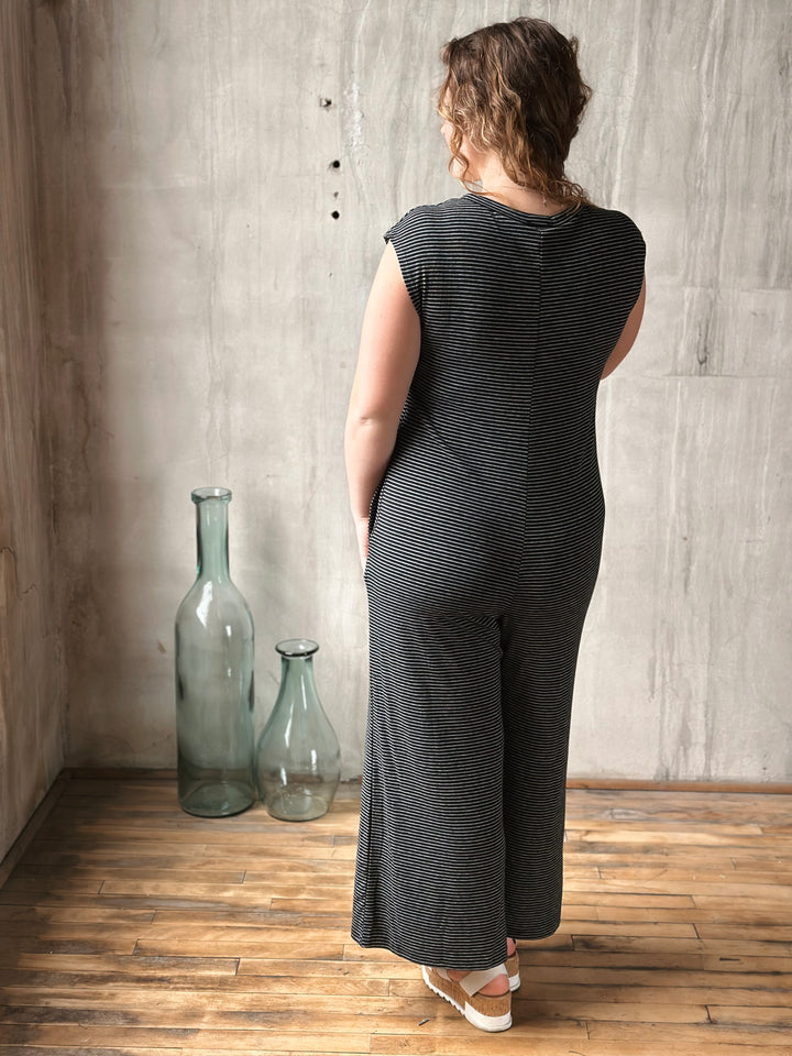 Person wearing a black and white striped sleeveless jumpsuit standing in a room with wooden flooring and a textured wall.