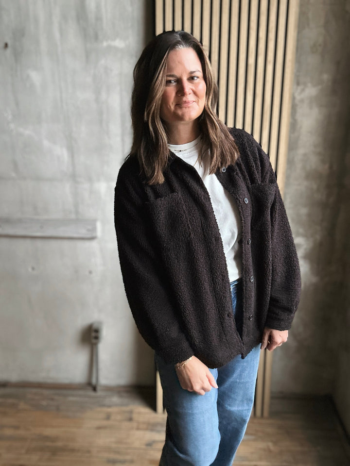 Woman wearing a black cardigan over a white shirt and blue jeans, standing against a concrete wall.