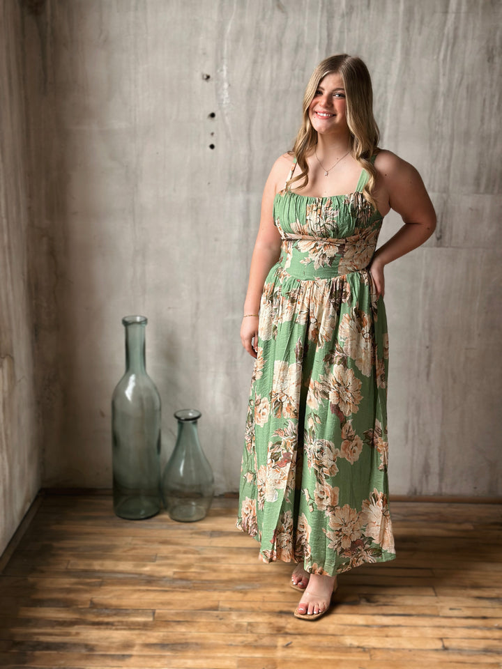 Woman wearing a green floral dress standing in a room with a textured wall and wooden floor.
