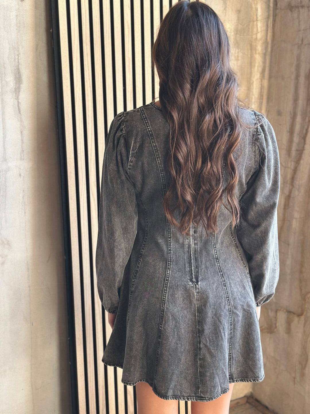 Person wearing a denim dress with long hair, standing against a neutral wall.