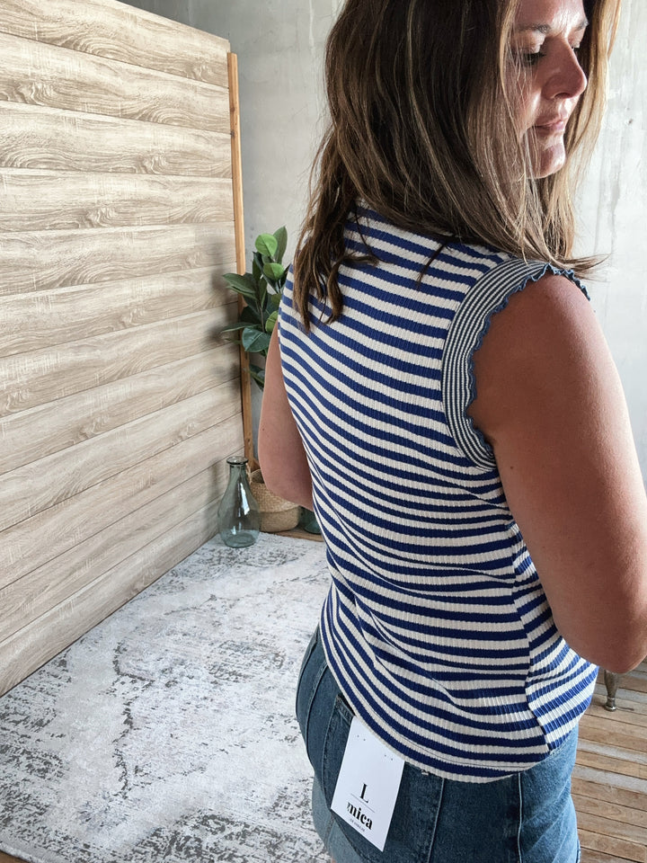 Woman wearing a blue and white striped sleeveless top standing in a room with wooden flooring and a textured wall.