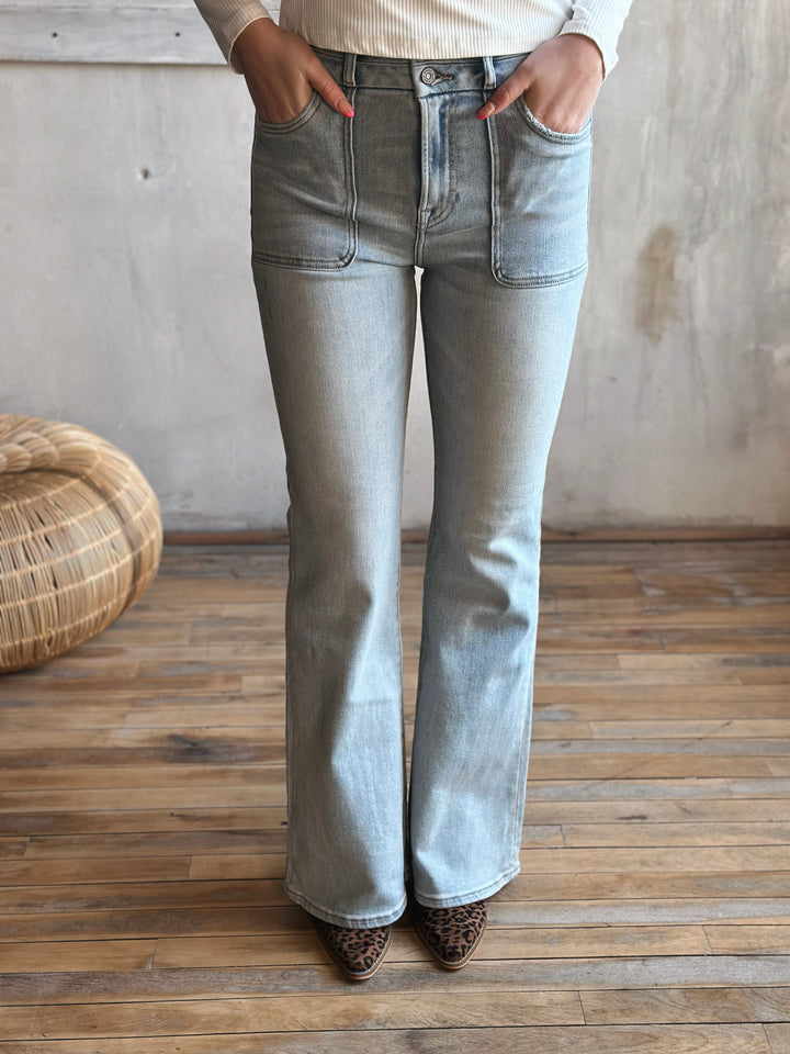 Person wearing light blue flared jeans standing on a wooden floor with a textured wall background.