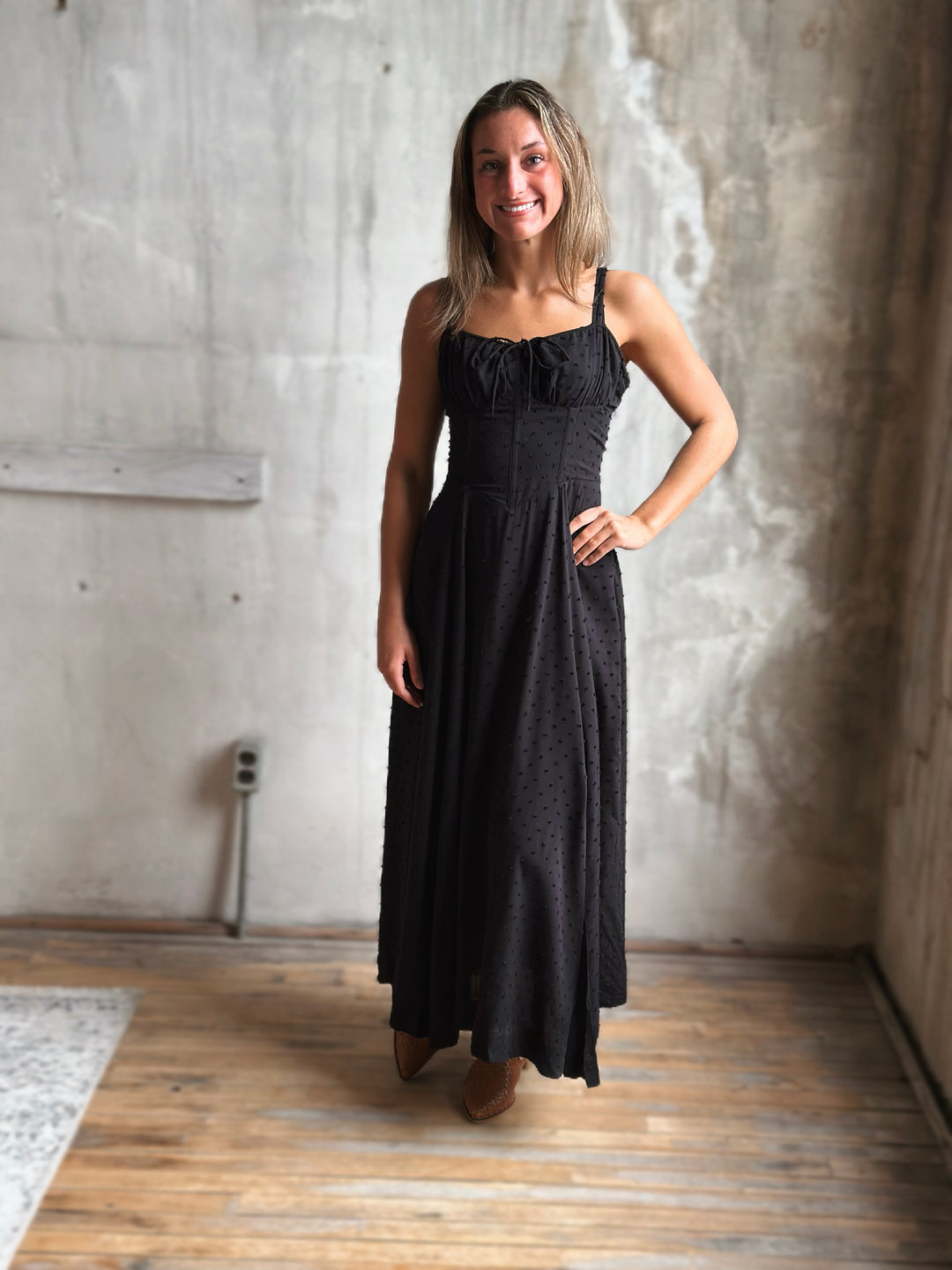 Woman wearing a black dress standing in a room with concrete walls and wooden floor.