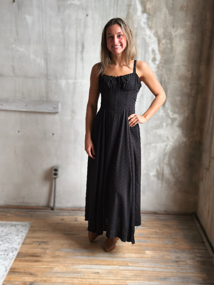 Woman wearing a black dress standing in a room with concrete walls and wooden floor.