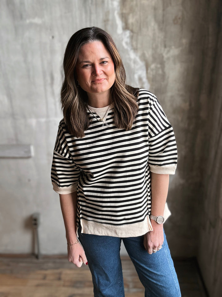 Woman wearing a black and white striped shirt and blue jeans standing against a textured wall.