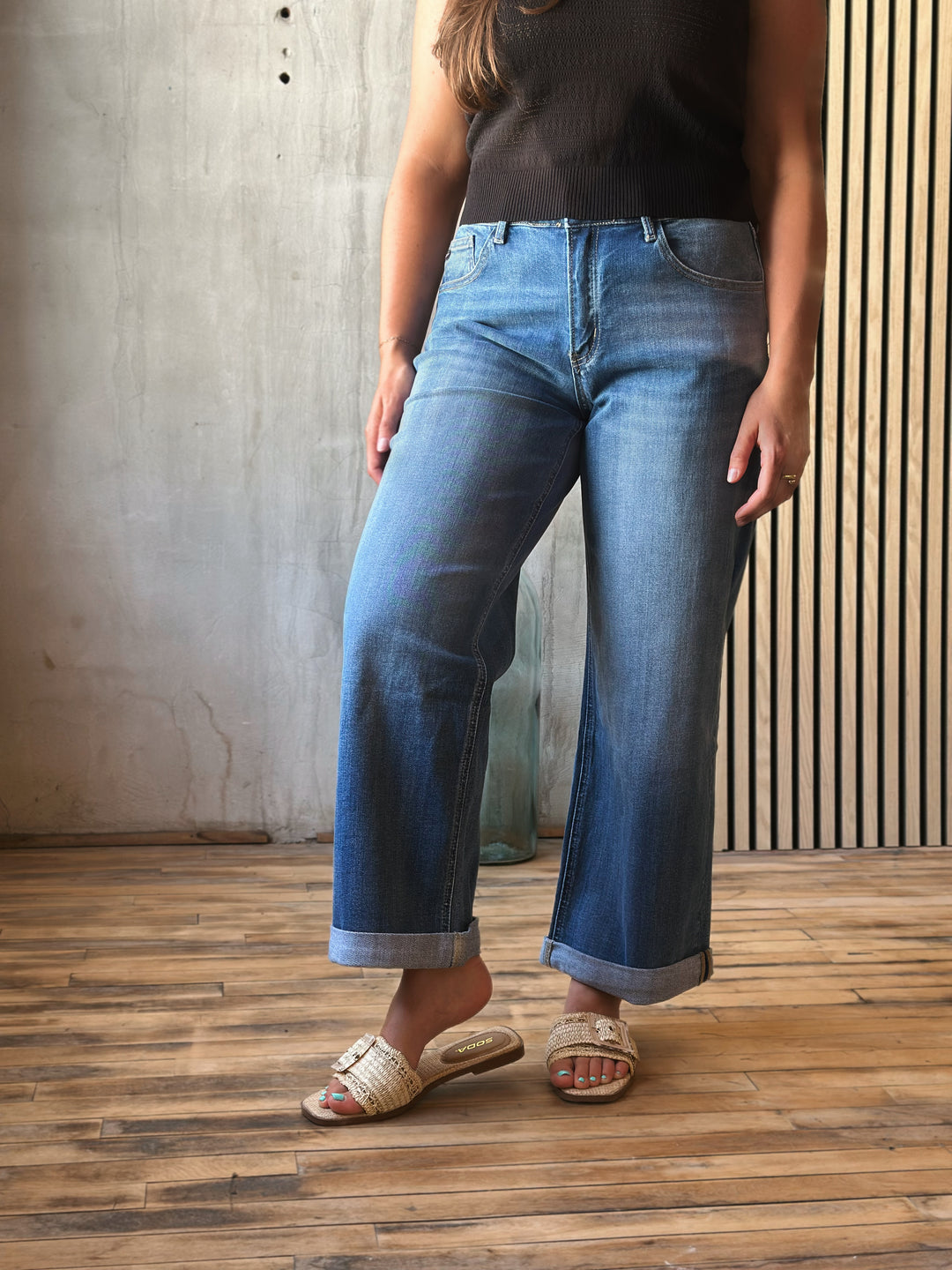 Person wearing blue jeans and a black top standing on a wooden floor with a concrete wall background.