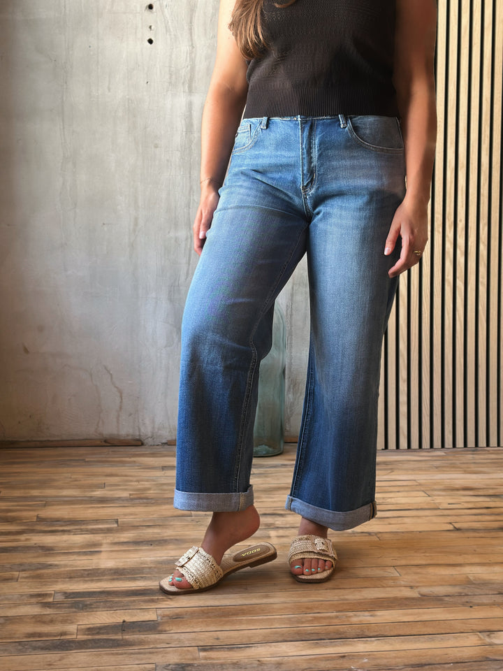 Person wearing blue jeans and a black top standing on a wooden floor with a concrete wall background.