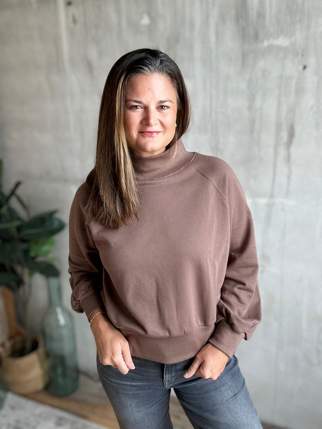 A woman wearing a brown mock neck sweatshirt and blue jeans, standing in front of a textured gray concrete wall with a plant in the background.