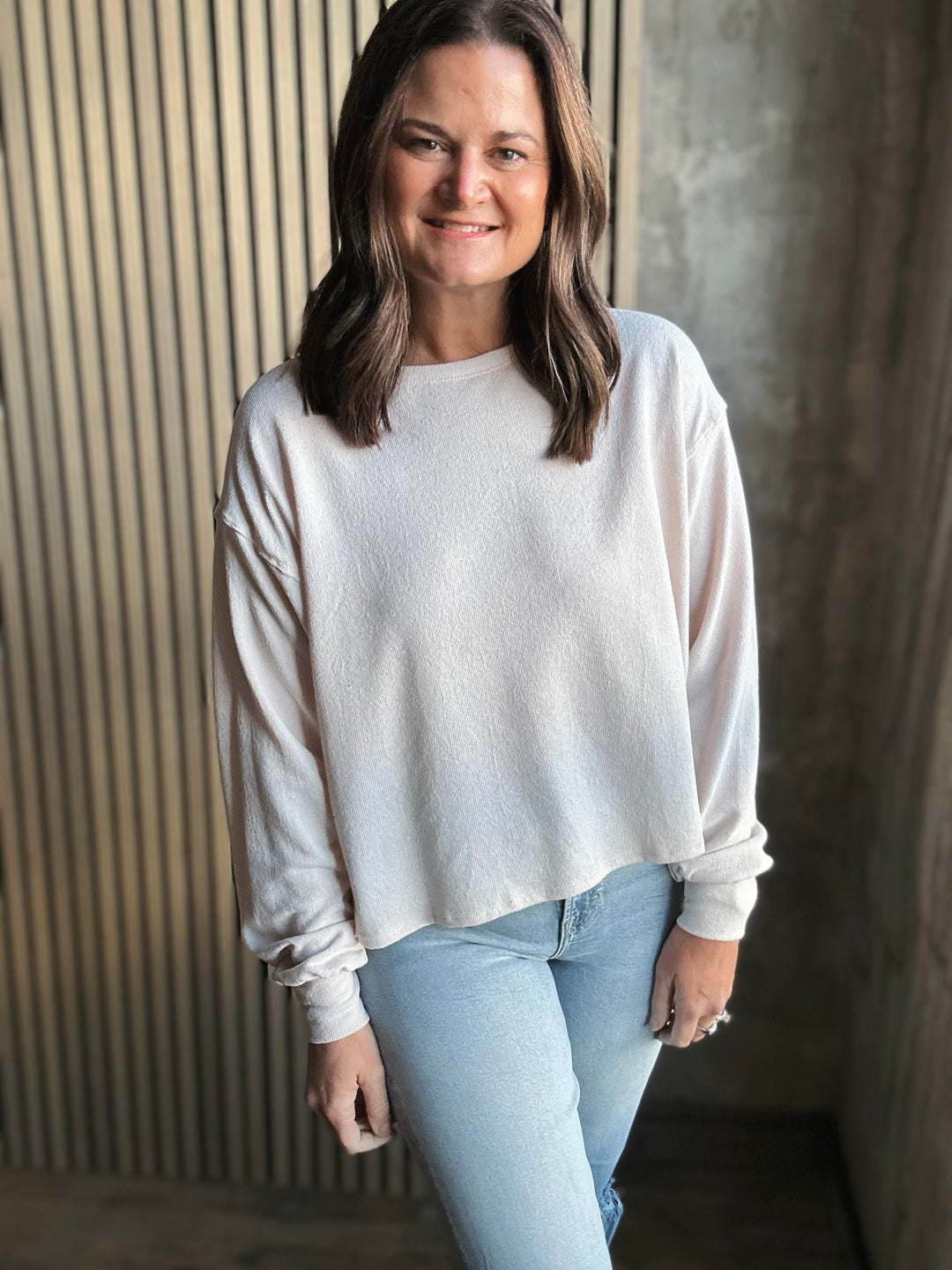 Woman wearing a white sweater and light blue jeans standing against a textured wall.