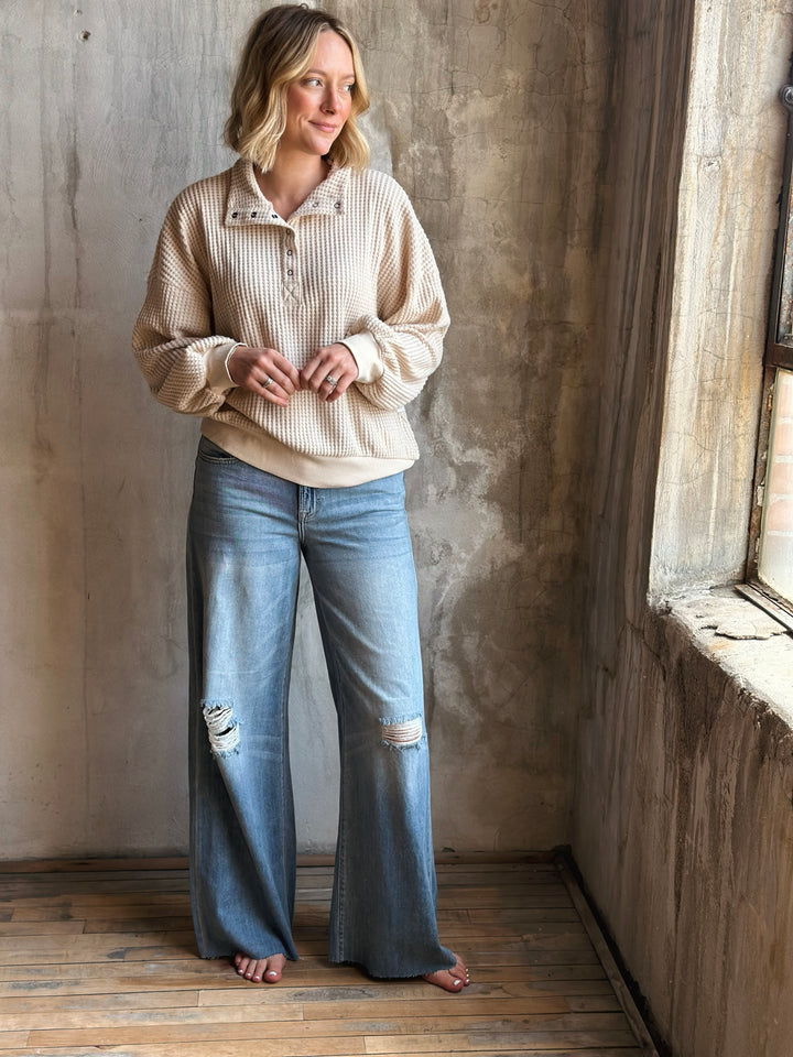 Woman wearing a beige sweater and blue jeans standing in a room with a textured wall and wooden floor.