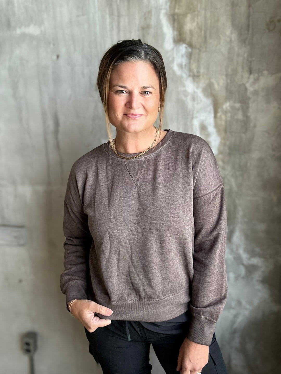 A woman wearing a heathered taupe-brown crewneck sweatshirt and black pants, standing against a textured concrete wall.