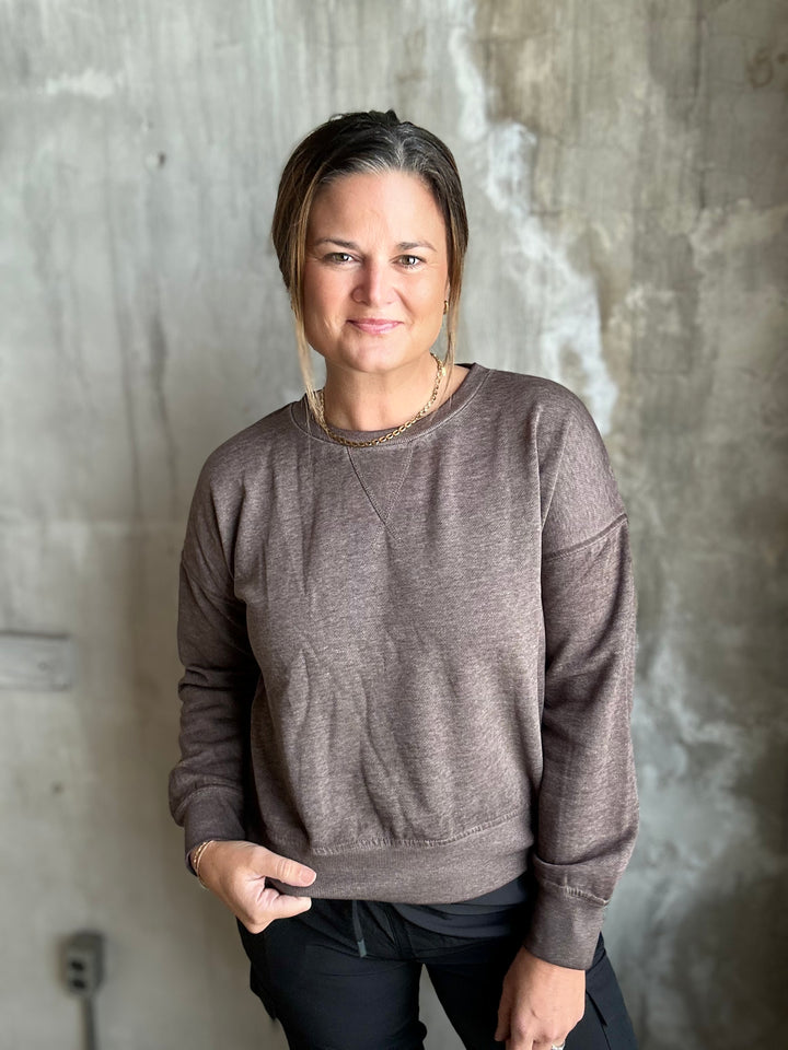 A woman wearing a heathered taupe-brown crewneck sweatshirt and black pants, standing against a textured concrete wall.