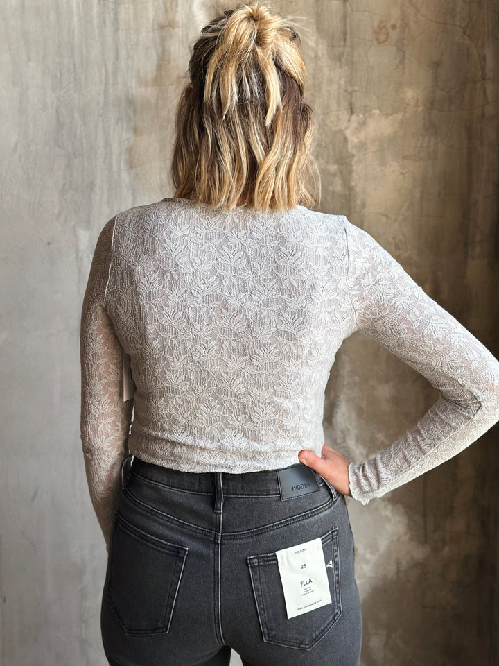 Person wearing a light gray long-sleeve top and dark jeans against a textured wall.