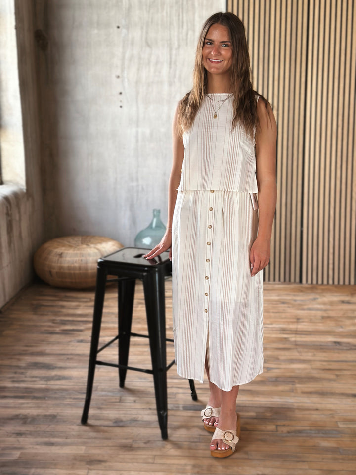 Woman in a white and beige outfit  standing in a room with a wooden floor and striped wall.